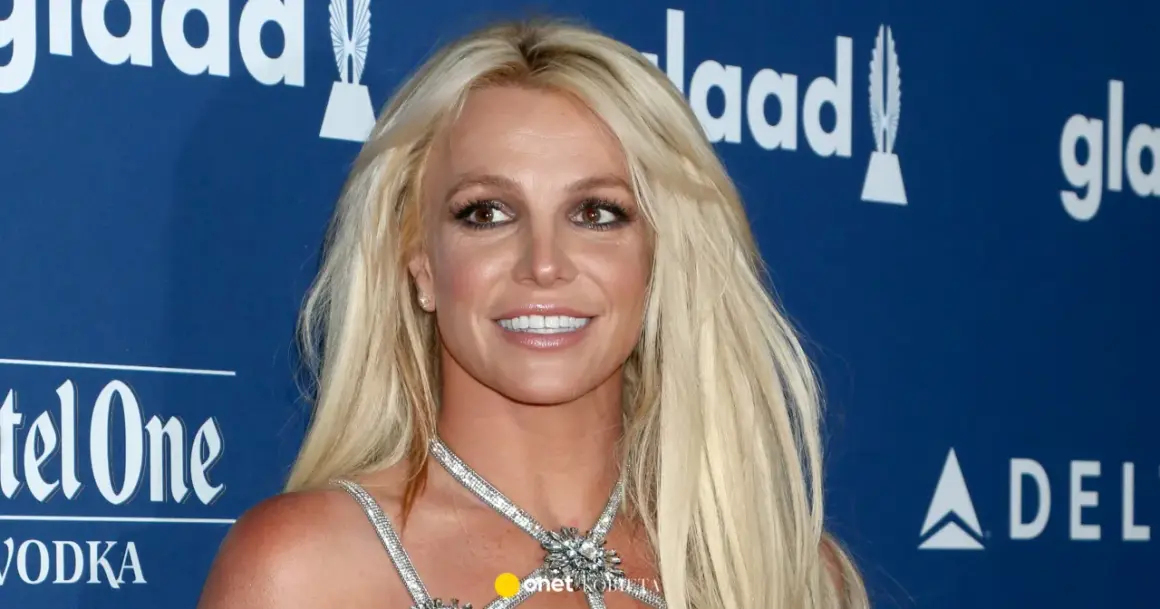 Uśmiechnięta Britney Spears, której wiek nie jest widoczny na zdjęciu, w błyszczącej kreacji na tle niebieskiej ścianki z logo GLAAD.