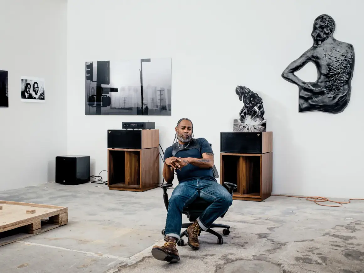 A man sits in an office chair in a gallery space, surrounded by art and audio equipment, a scene that feels like a snapshot of Arthur Jafa's creative process.