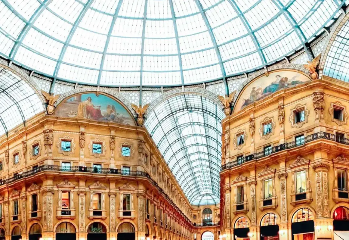 Inside Milan's Galleria Vittorio Emanuele II, a stunning glass dome arches over ornate buildings adorned with frescoes, reminiscent of grand Milan art galleries.