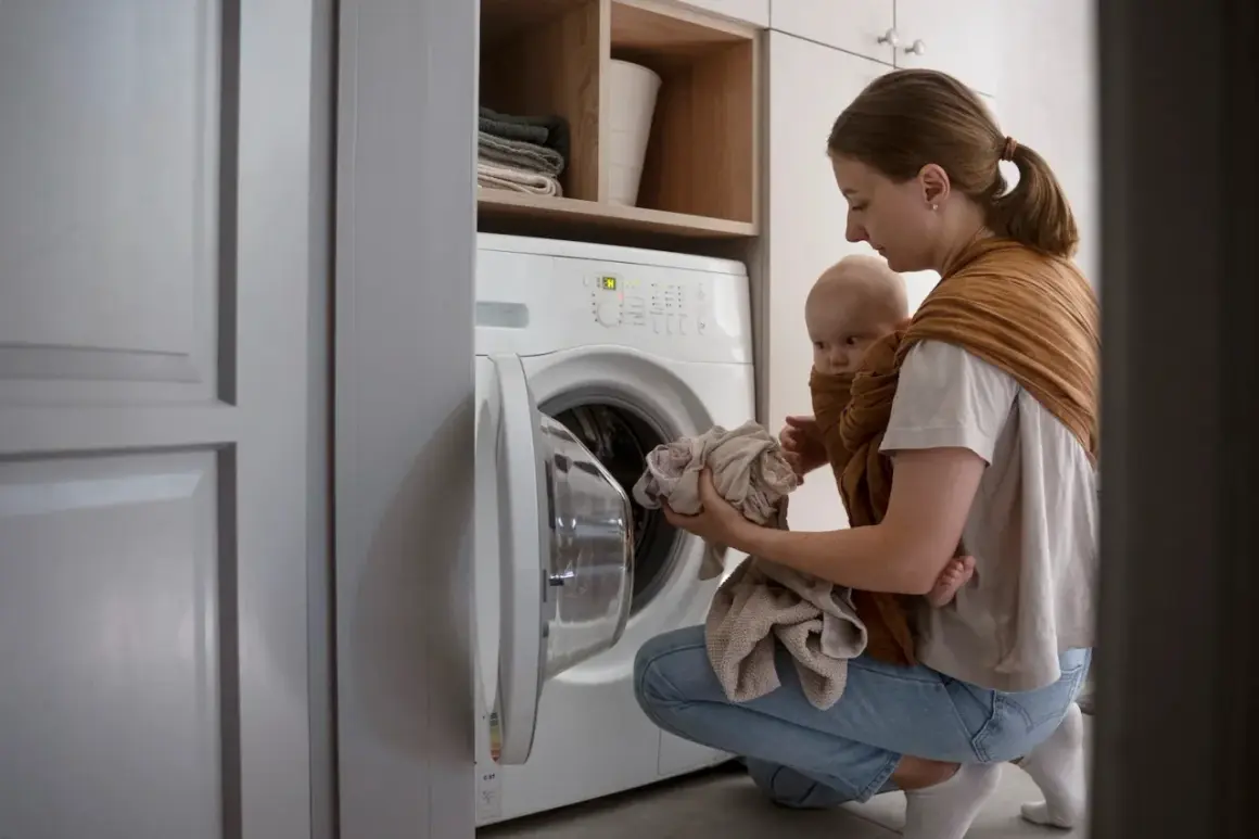 Une mère porte son bébé en écharpe tout en chargeant le linge dans le lave-linge. Une astuce de grand-mère pour éviter la mauvaise odeur du lave-linge.