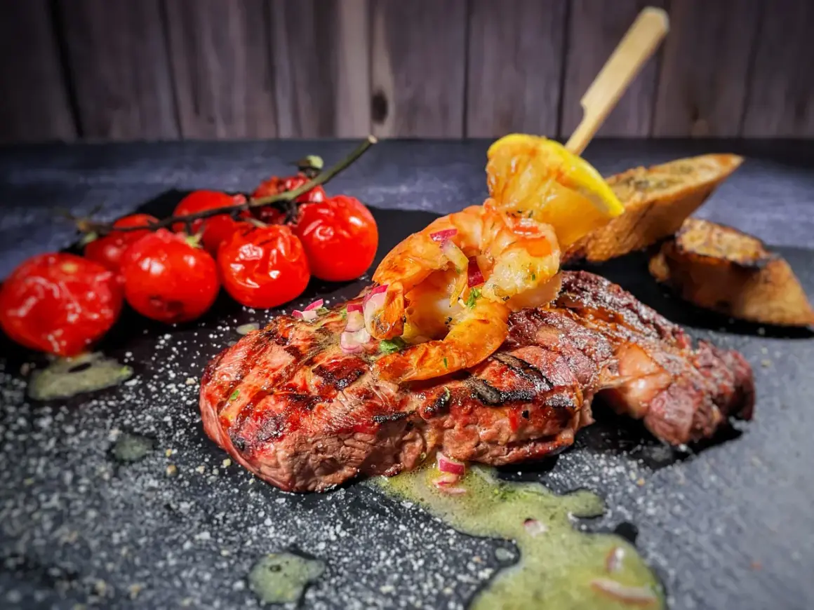Perfektes Surf and Turf Rezept: saftiges Steak mit gegrillten Garnelen, Kirschtomaten und Zitronenspalte auf schwarzem Stein.