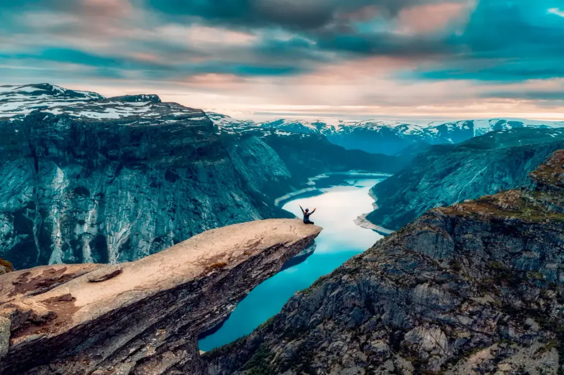 En person sitter på Trolltungan i Norge, med armarna utsträckta mot den dramatiska fjällnaturen och en glittrande sjö nedanför.