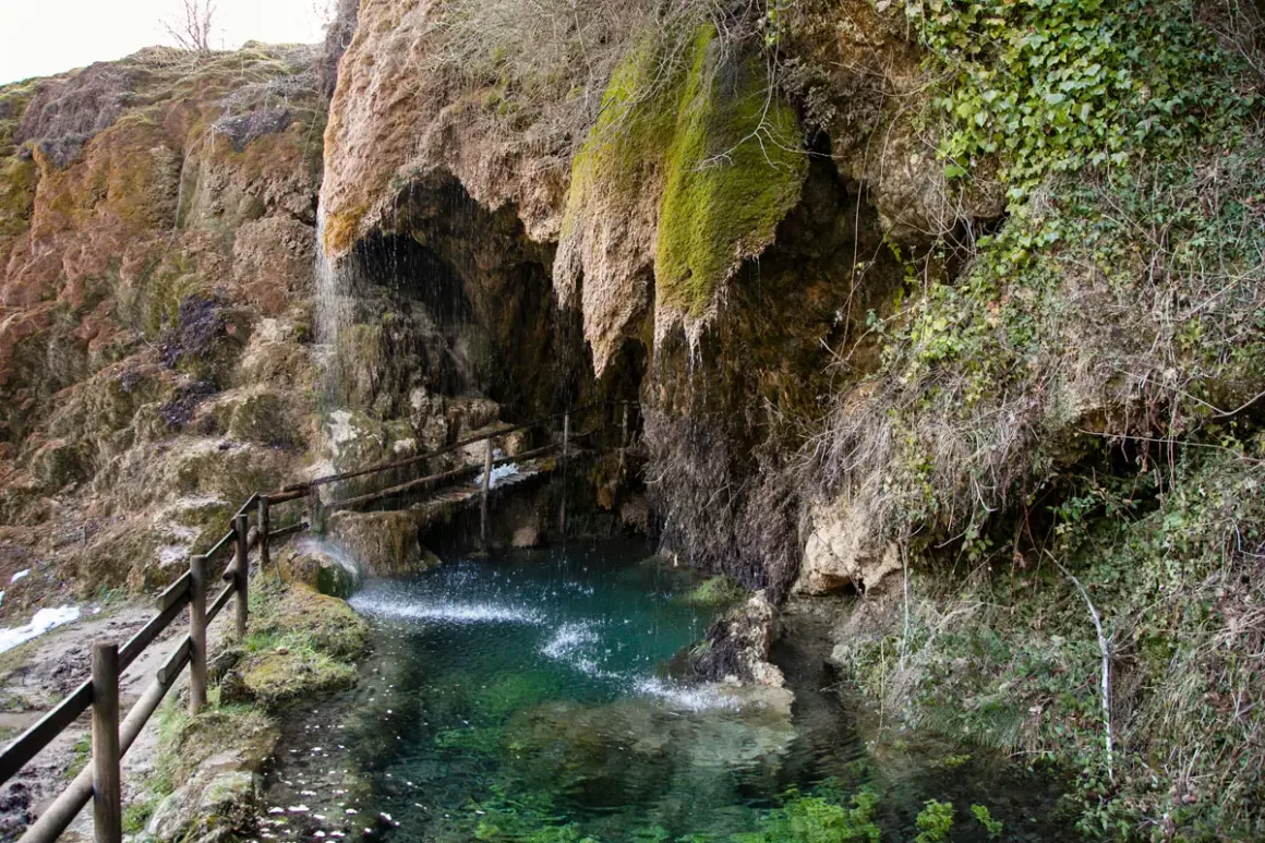 Cascata che scende tra rocce muschiose, formando una pozza d'acqua cristallina. Un sentiero con staccionata in legno conduce alle grotte di Labante.