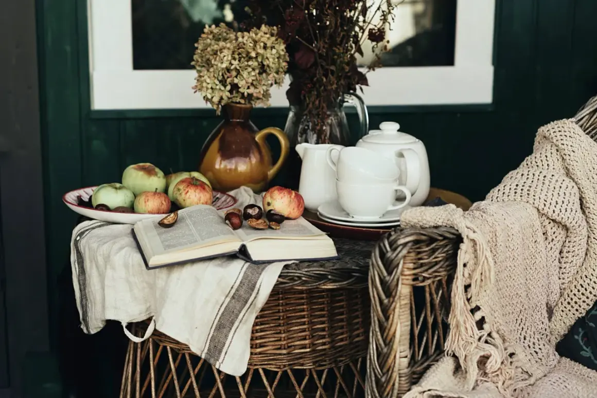 A cozy scene with apples, chestnuts, and an open book on a linen cloth, embodying the cottagecore aesthetic. A teapot and cups sit nearby, with a knitted blanket draped over a wicker chair.