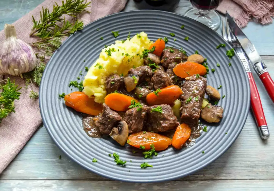 Herzhaftes Boeuf Bourguignon Rezept: zartes Rindfleisch mit Karotten, Pilzen und Kartoffelpüree auf einem grauen Teller, garniert mit Petersilie.