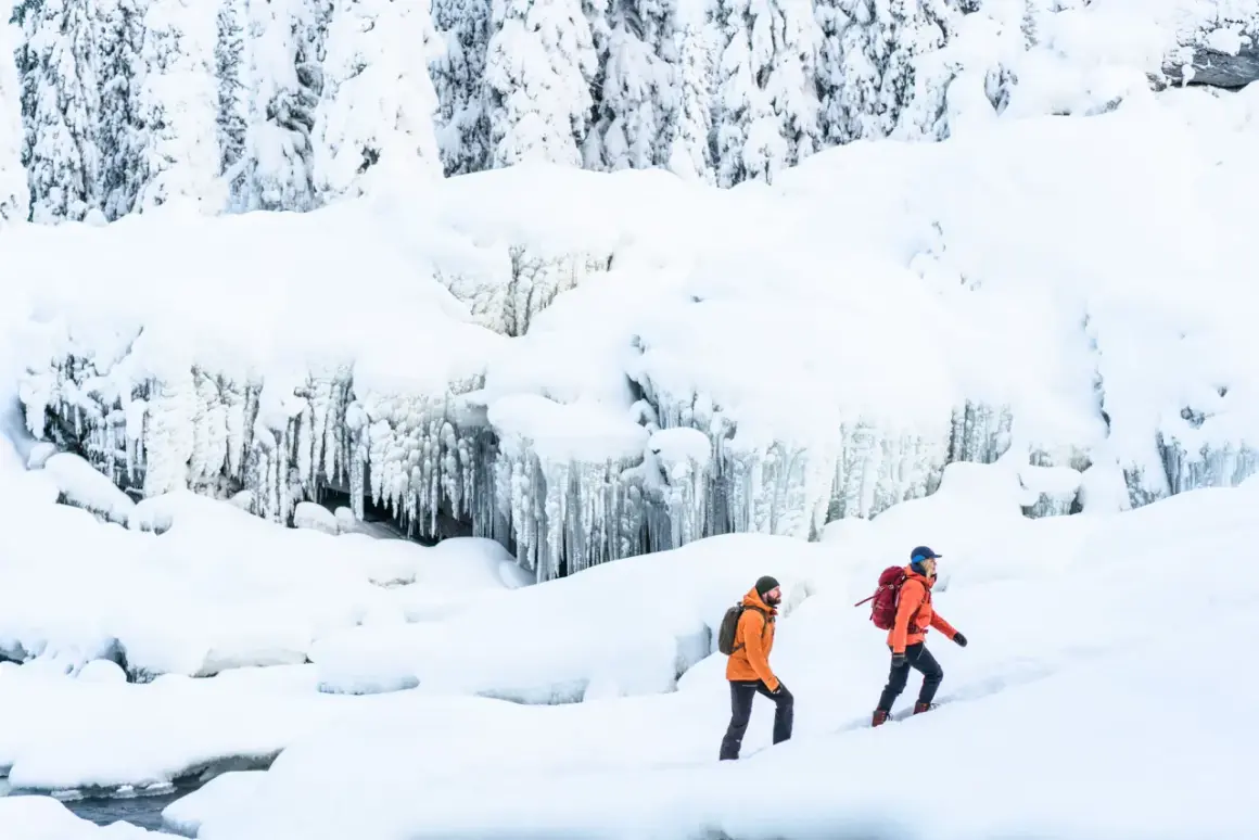 Två personer med ryggsäckar vandrar på vintern i ett snöigt landskap med istappar.