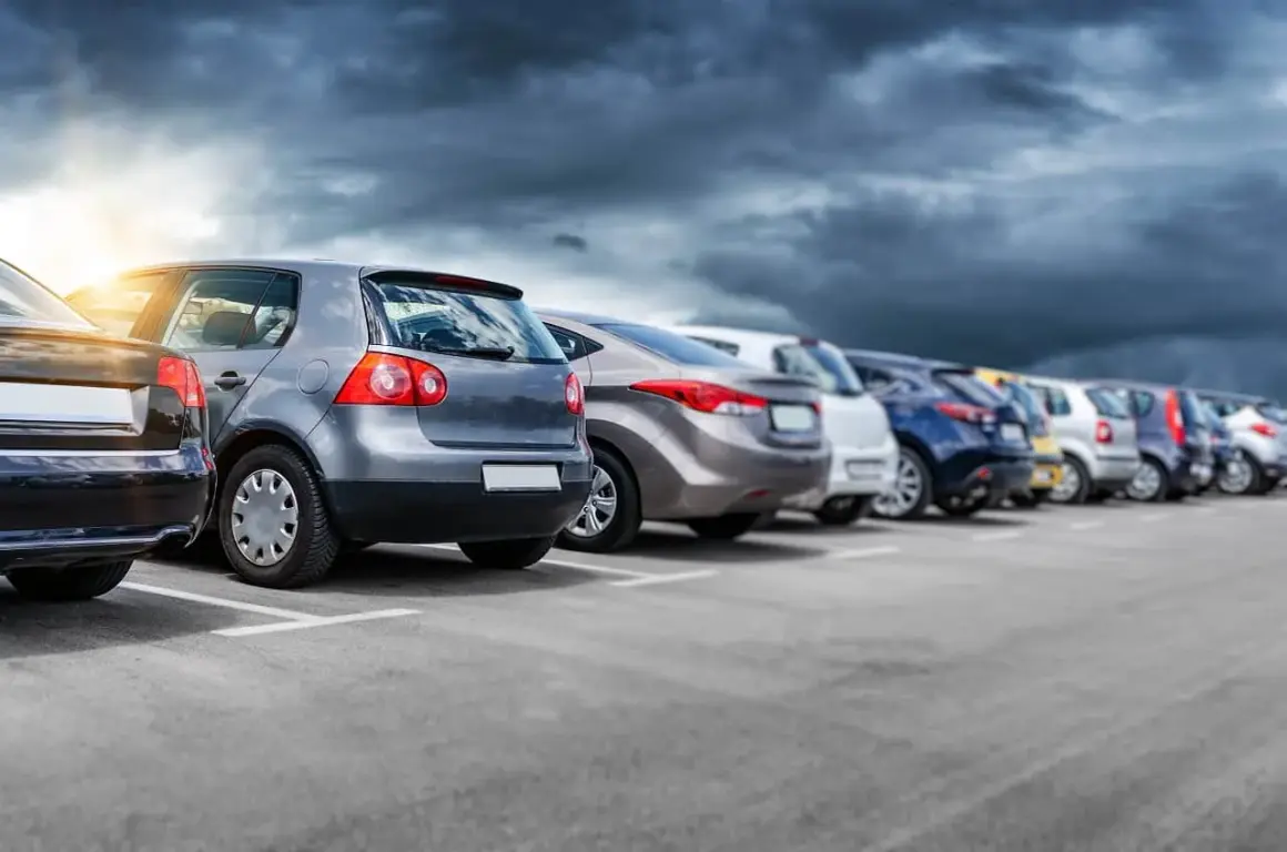 Rząd samochodów na parkingu, w tym jeden z włoską rejestracją, pod burzowym niebem.