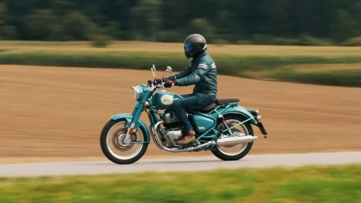 Mężczyzna w kasku i skórzanej kurtce jedzie turkusowym Royal Enfield Classic 650 po wiejskiej drodze, otoczonej polami.