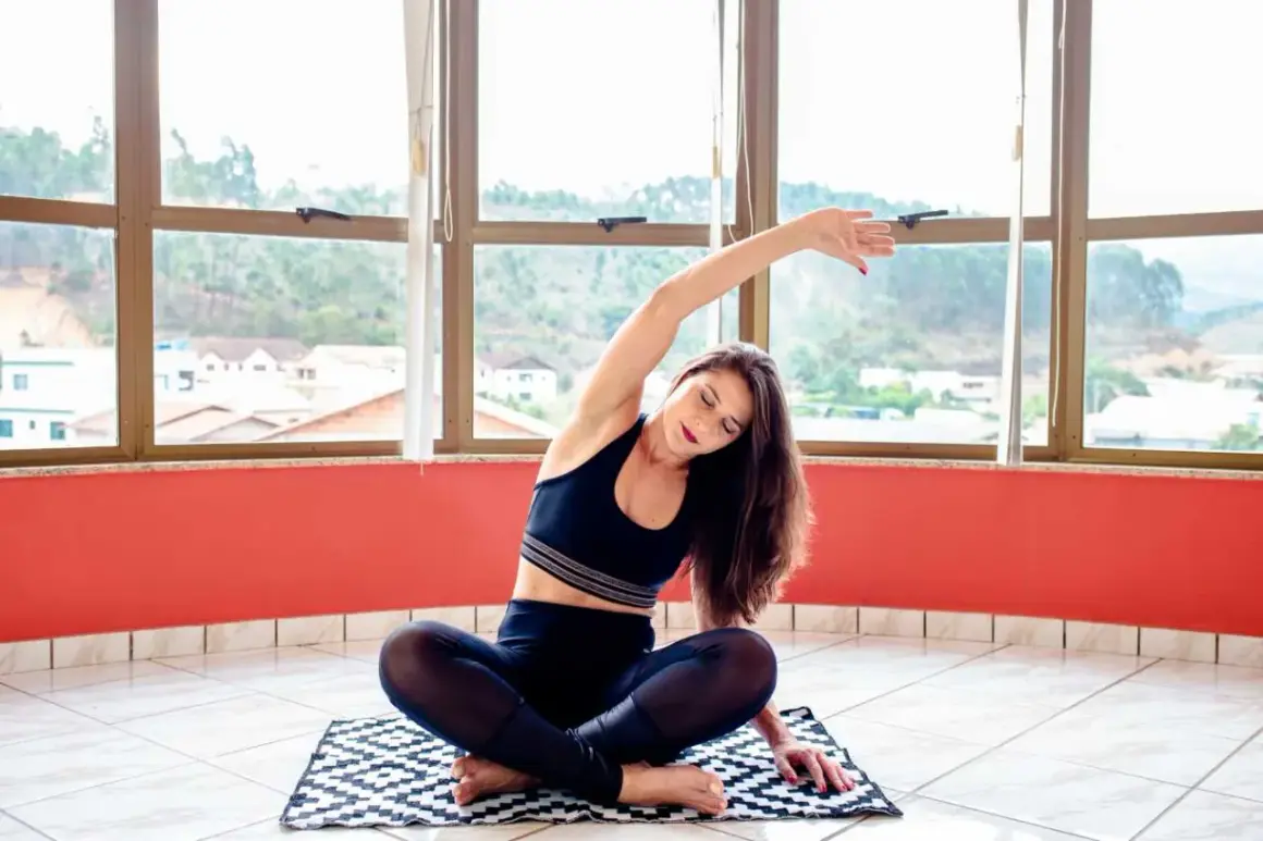 Femme pratiquant des exercices de rééducation périnée à la maison, assise en tailleur, étirant son corps près d'une grande fenêtre.