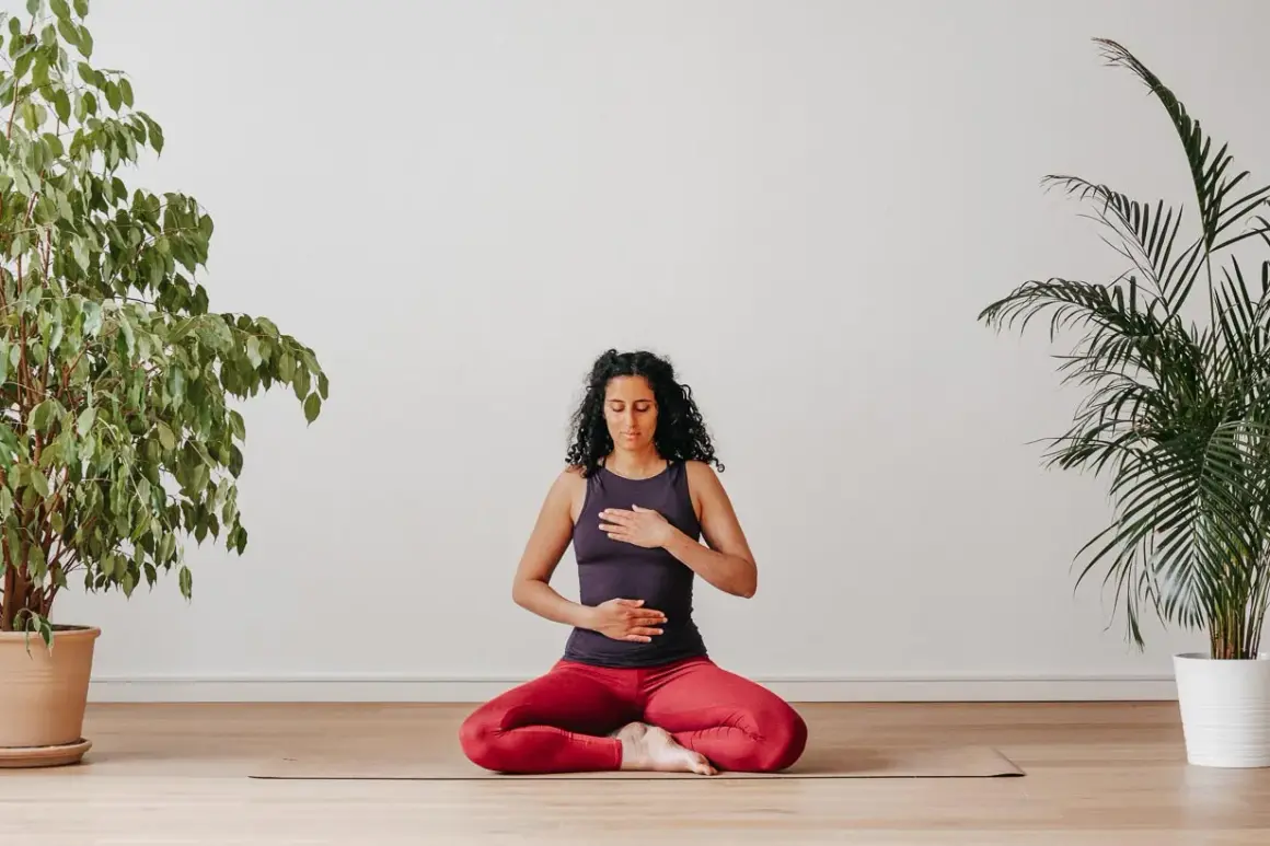 Frau sitzt im Schneidersitz auf einer Yogamatte, Hände auf Brust und Bauch. Perfekt, um mit Yoga anzufangen und innere Ruhe zu finden.