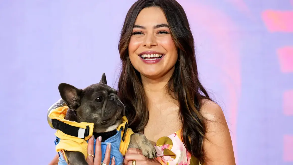 Miranda Cosgrove lächelt und hält einen französischen Bulldoggen-Welpen in einem gelben Outfit. Vielleicht ein neuer Co-Star für ihre Filme?