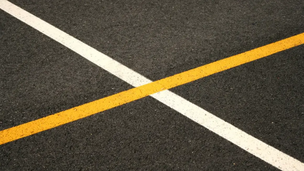 Nowe malowanie pasów drogowych: biała i żółta linia przecinają się na ciemnym asfalcie, tworząc geometryczny wzór.