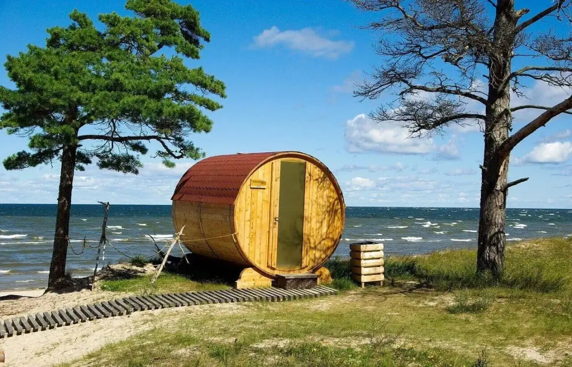 Beczka-sauna nad morzem, jedna z nietypowych atrakcji Juraty. Drewniana konstrukcja z czerwonym dachem, przy plaży, pod drzewami.