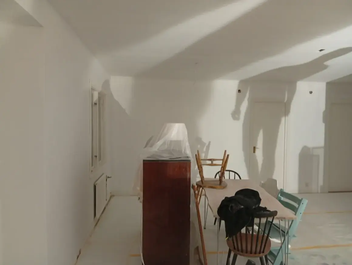 Rummet är nymålat vitt, möbler är täckta med plast. Skuggor på väggarna antyder att det är en pågående renovering, vilket väcker frågan: hur lång tid tar det att måla ett rum?