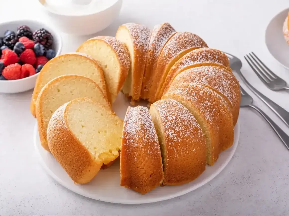 Pyszna babka piaskowa, jak z przepisu babci, posypana cukrem pudrem, z owocami i widelcami obok.