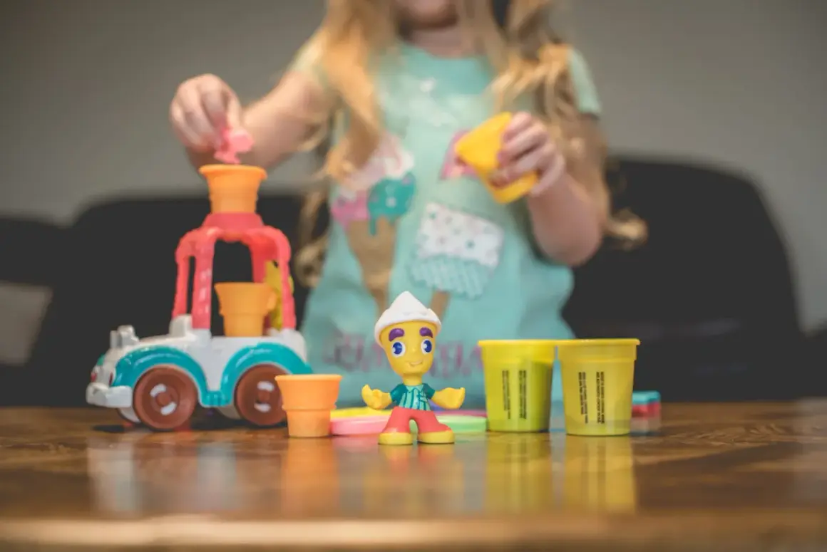 Dziecko bawi się w play doh warsztat, tworząc lodowe przysmaki. Na stole kolorowe foremki i figurka lodziarza.