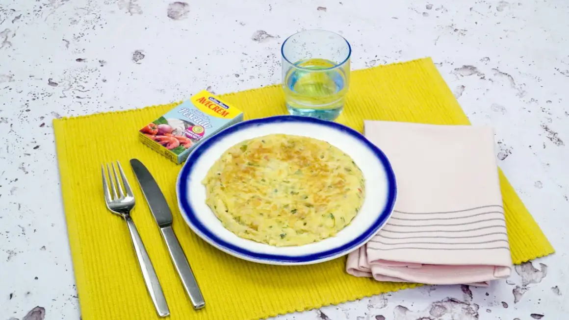 Tortilla de gambas dorada en plato blanco con borde azul, servida con cubiertos, vaso de agua y caldo de pescado.