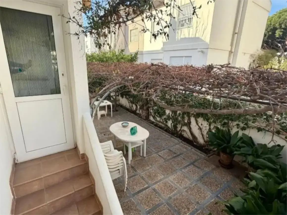 Patio con mesa y sillas bajo una pérgola cubierta de enredaderas secas. Un gran cortijo se vislumbra al fondo.