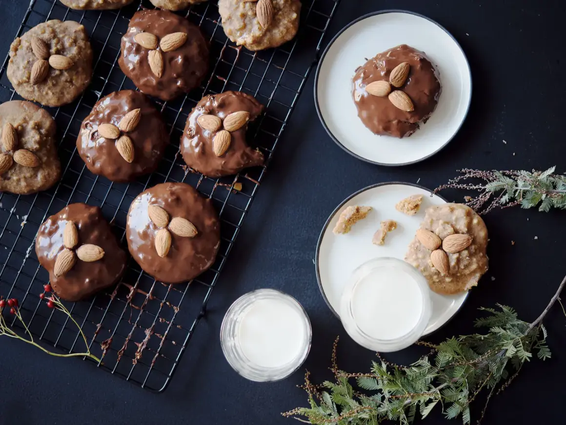 Schokoladenüberzogene Elisenlebkuchen mit Marzipan und Mandeln auf einem Gitter, daneben Milchgläser und Tannenzweige.