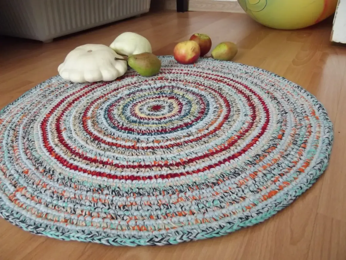 Tappeto uncinetto rotondo con fantasie a spirale multicolore. Sopra, zucche bianche, una pera e mele rosse.