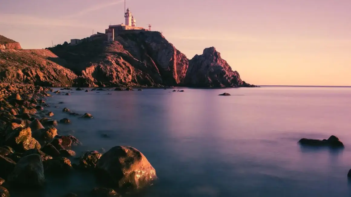 Faro en un acantilado rocoso al atardecer, con el mar tranquilo y piedras en la orilla. Parece la boca de los frailes.