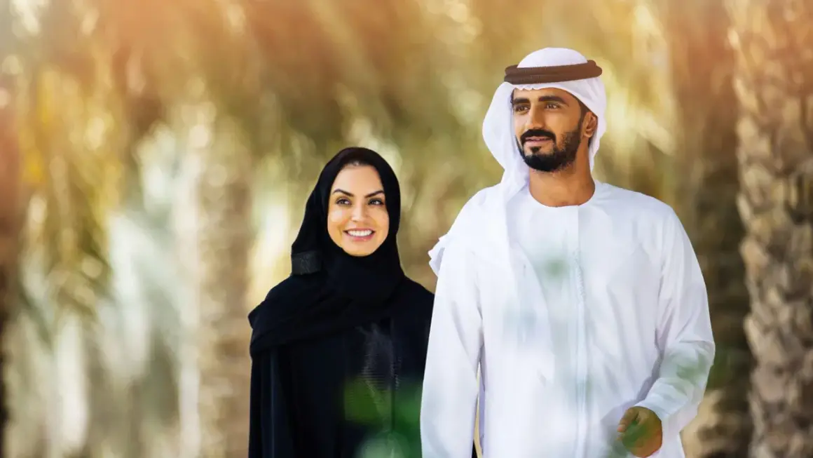 Uśmiechnięta kobieta w tradycyjnym stroju arabskim damskim i mężczyzna w białej dżalabiji spacerują wśród palm.