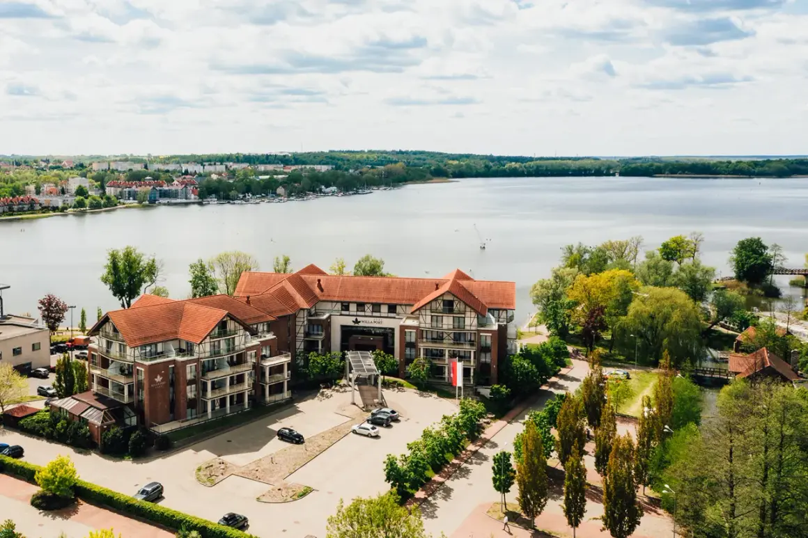 Willa Port w Ostródzie, hotel z widokiem na jezioro, otoczony zielenią i zabudowaniami.