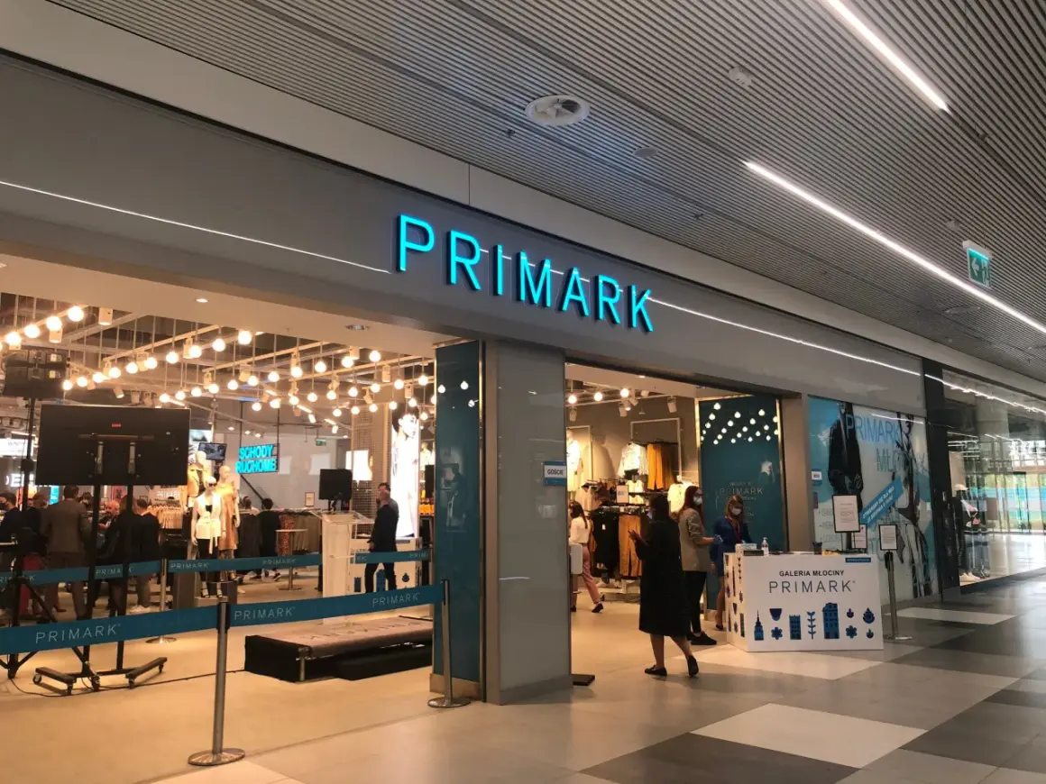 Wejście do sklepu Primark. W środku widać manekiny i ubrania. Na ladzie informacyjnej znajduje się **Primark tabela rozmiarów**.