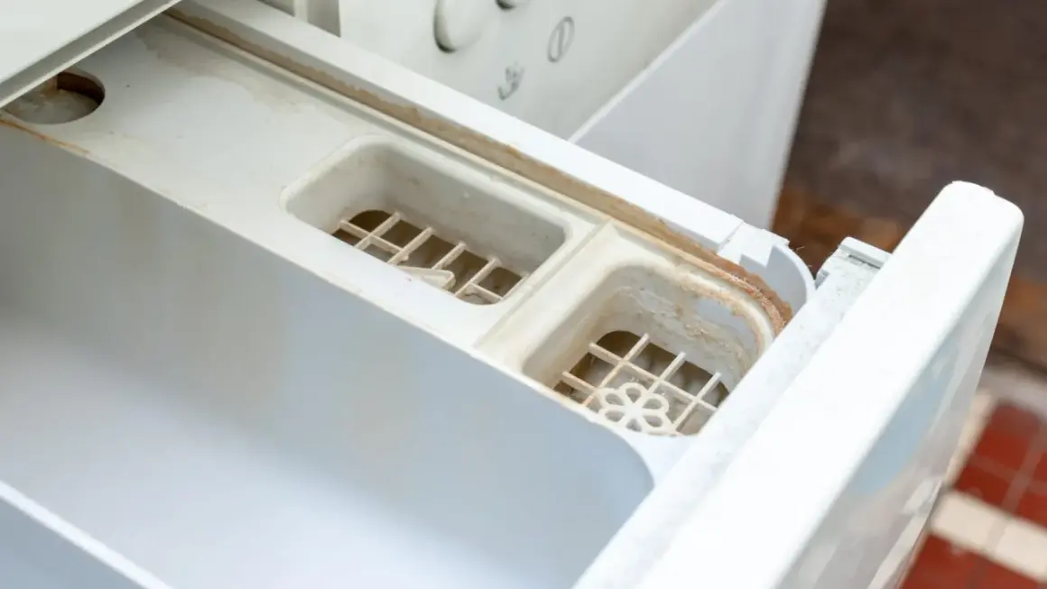 Schmutziges Waschmittelfach einer Waschmaschine, das dringend eine Reinigung benötigt. Kalk und Seifenreste haben sich abgelagert.