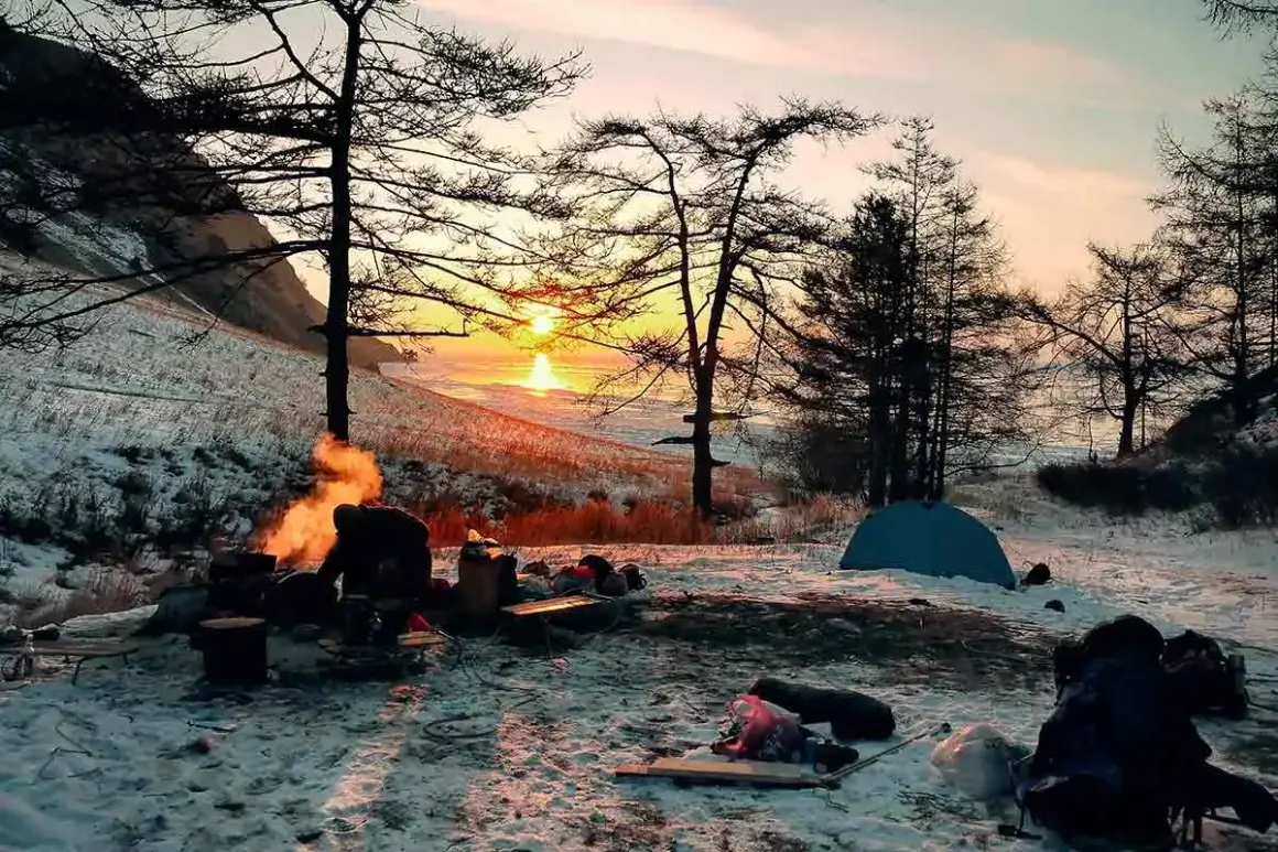 Campamento invernal con fogata y tienda de campaña. La puesta de sol ilumina el paisaje nevado, un ejemplo de supervivencia y naturaleza.