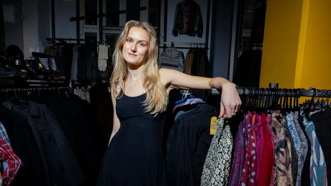 En ung kvinna i en svart klänning lutar sig mot en klädställning med färgglada kläder i en butik. Kanske hittar hon nya fynd för sitt friis company.