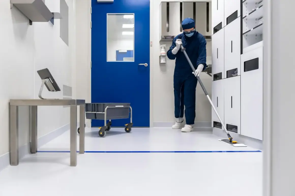 Osoba w kombinezonie i masce myje podłogę w sterylnym, clean room. W tle niebieskie drzwi i białe szafki.