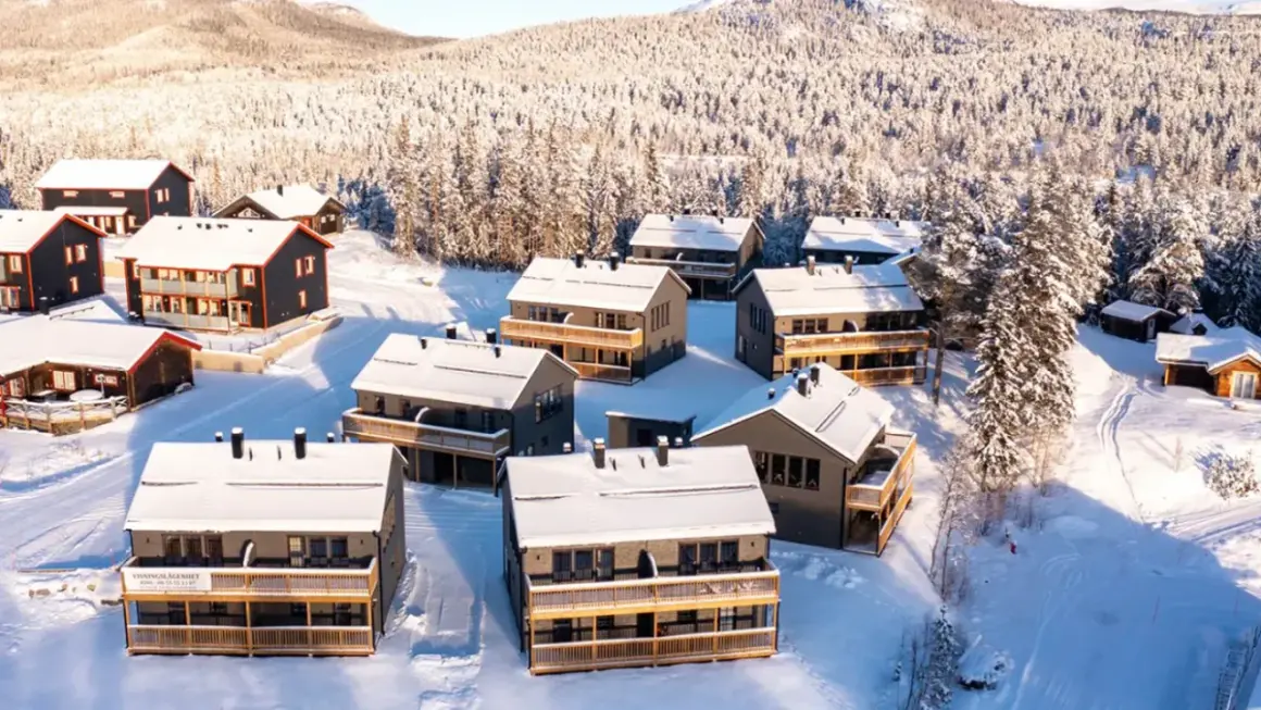 Vinterlandskap med snötäckta hus i Funäsdalens stugby. Solen lyser på de moderna stugorna och den snöklädda skogen.