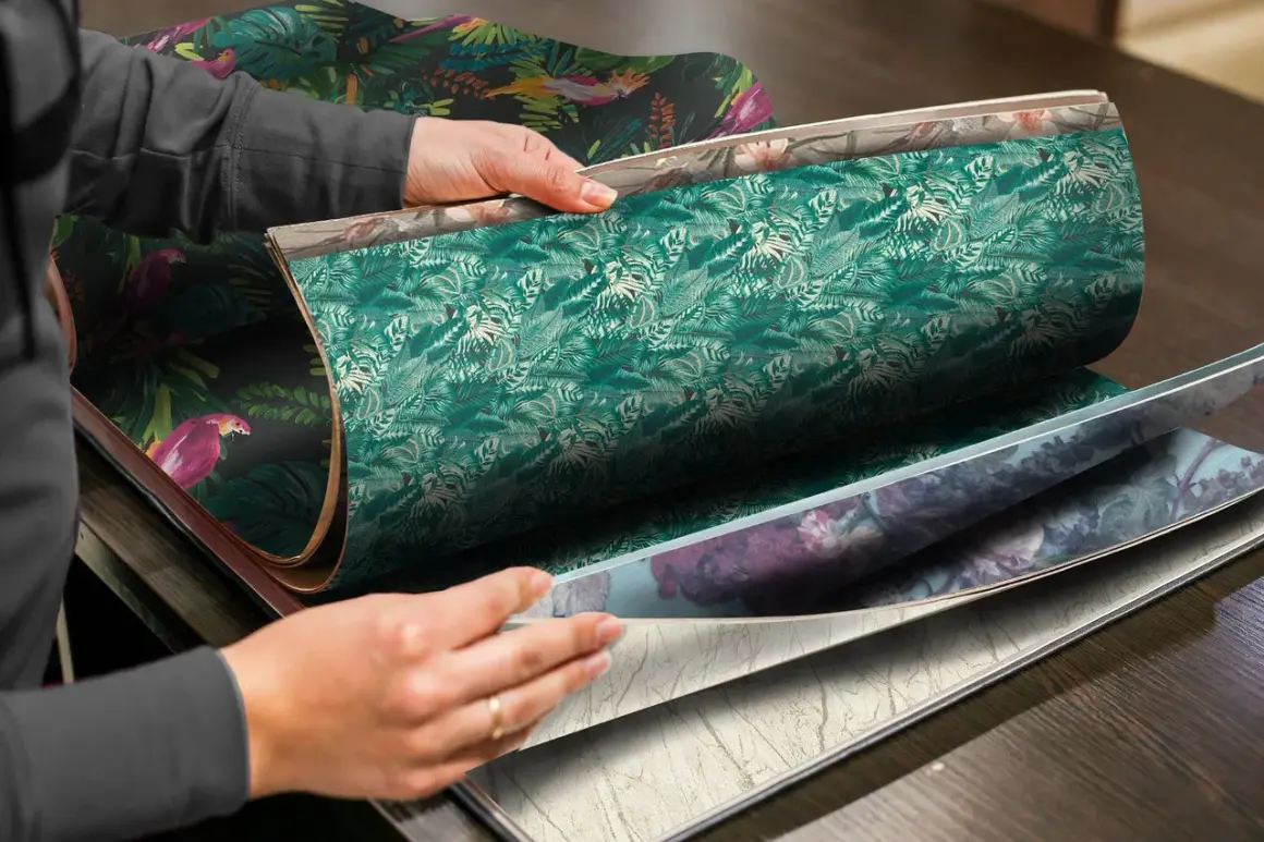 Händer bläddrar i en tapetbok med djungelmotiv och blomstermönster. Perfekt för att välja non woven lim till papperstapet.