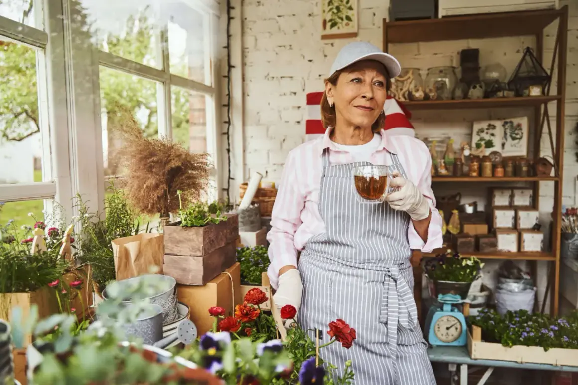 Frau mit Kappe und Schürze trinkt Tee im Blumenladen. Sie genießt ihre Zeit, vielleicht im Rahmen von altersteilzeit modellen, umgeben von Pflanzen.