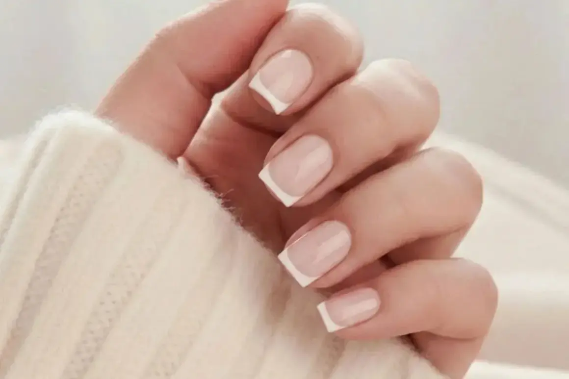 En hand med klassisk fransk manikyr på korta naglar, insvept i en mjuk, beige tröja.
