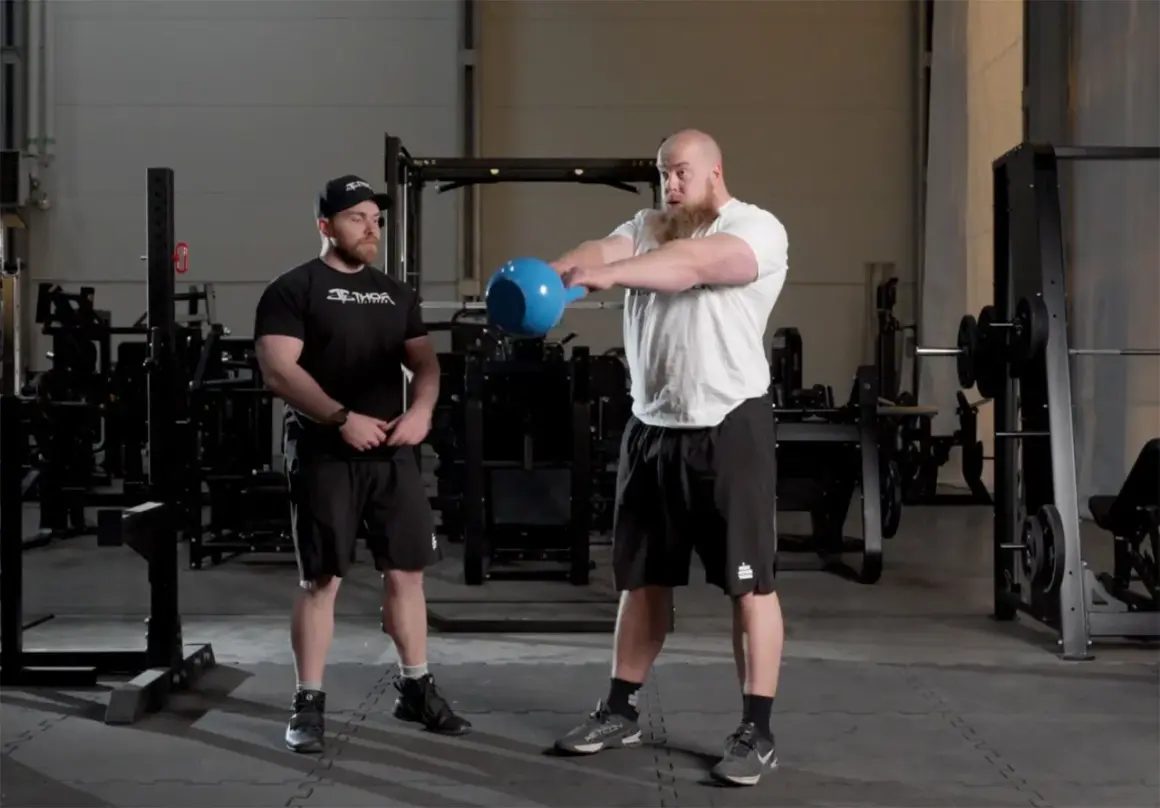 Två män i gym utför kettlebell swing. Den ena mannen, med skägg, håller i en blå kettlebell och är i rörelse. Den andra mannen står bredvid och observerar.
