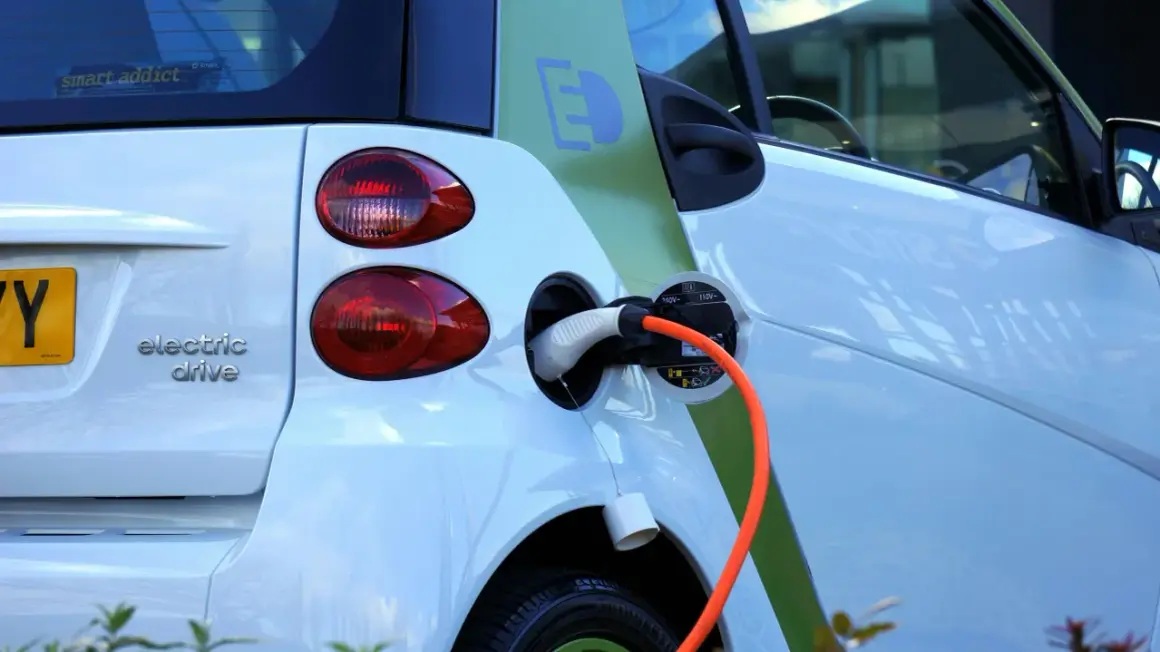 Voiture électrique blanche en charge, avec un câble orange branché. Le symbole "E" vert sur le flanc indique son caractère écologique. Le frais réel d'une voiture électrique est sa recharge.