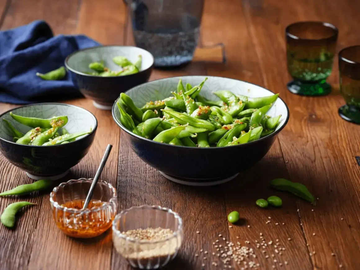 Schon gewusst, was ist Edamame? Grüne Sojabohnen in Schoten, gewürzt mit Sesam und Chili, serviert in Schalen auf Holztisch.