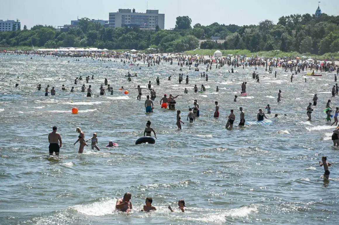 Tłum ludzi kąpiących się w Bałtyku, na tle plaży z parasolami i budynków.