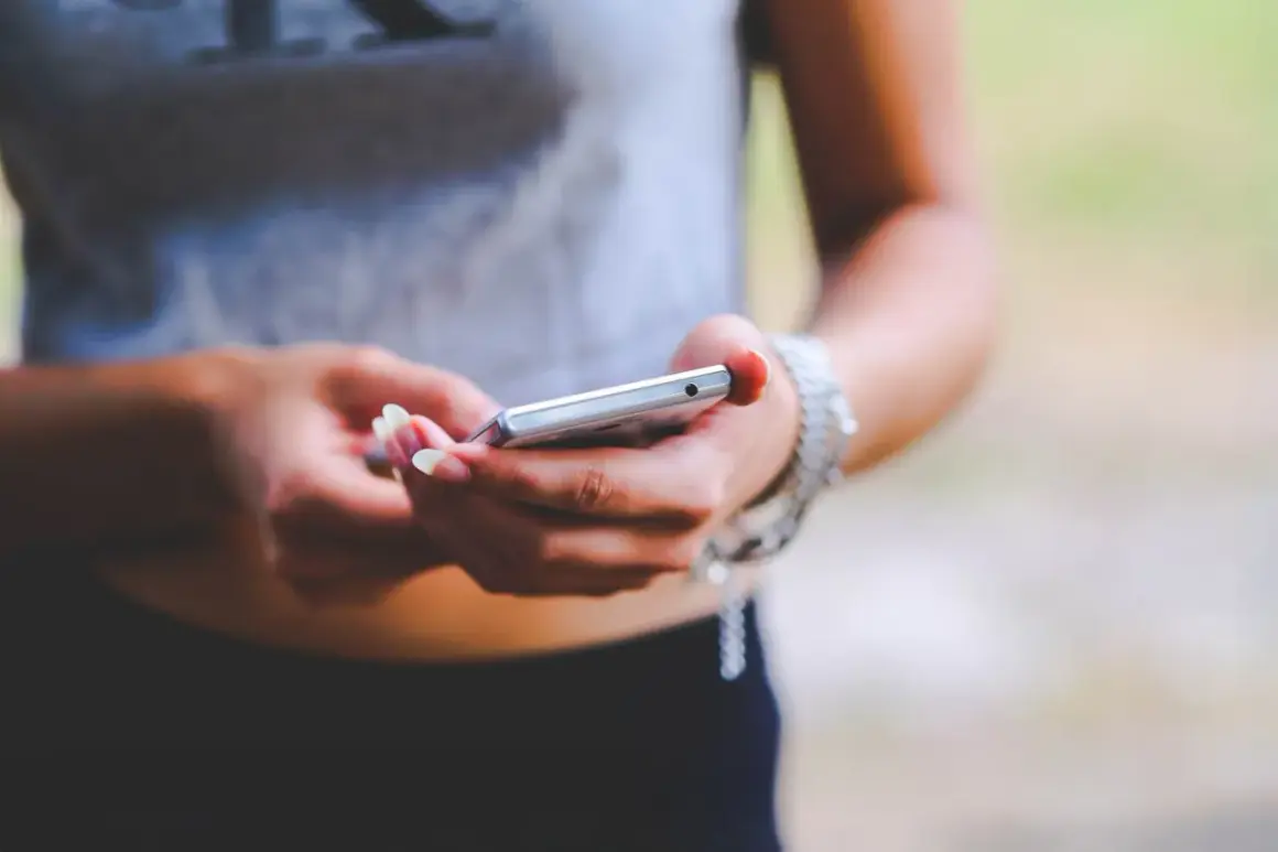 Dłonie z zadbanymi paznokciami trzymają srebrny telefon. To zrzut ekranu Samsunga, uchwycony w letnim otoczeniu.