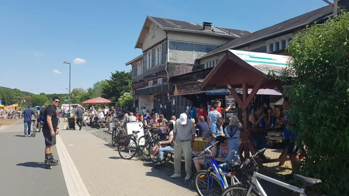 Menschen genießen das Leben am **Wuppertaler Hauptbahnhof**. Viele sitzen draußen, einige mit Fahrrädern, bei Sonnenschein.