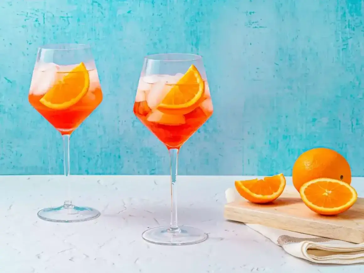 Två glas med Aperol Spritz, is och apelsinskivor, med en hel apelsin och skivor på en skärbräda i bakgrunden.