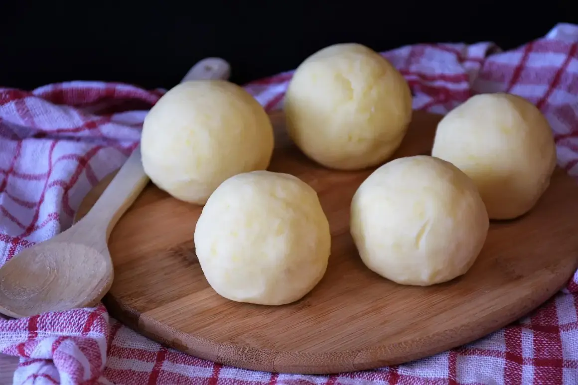 Pięć okrągłych, jasnych klusek na desce, obok drewniana łyżka. Czy można zrobić kluski śląskie z mąki pszennej?