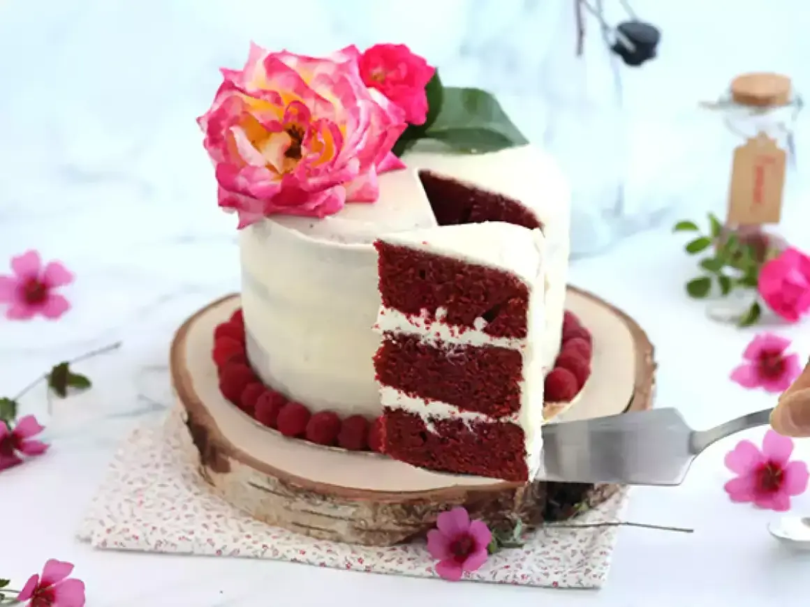 En vacker red velvet cake med fluffig frosting, dekorerad med hallon och rosa blommor. En bit skärs upp.
