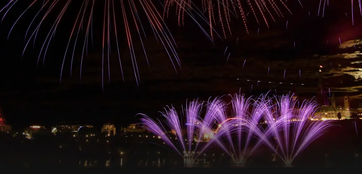 Feu d'artifice violet et blanc illuminant un ciel nocturne au-dessus d'une ville et de bâtiments historiques.