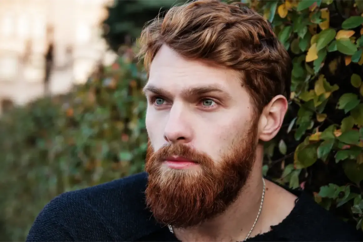Un homme au visage ovale, avec une barbe rousse et des yeux bleus perçants, regarde au loin, entouré de verdure.