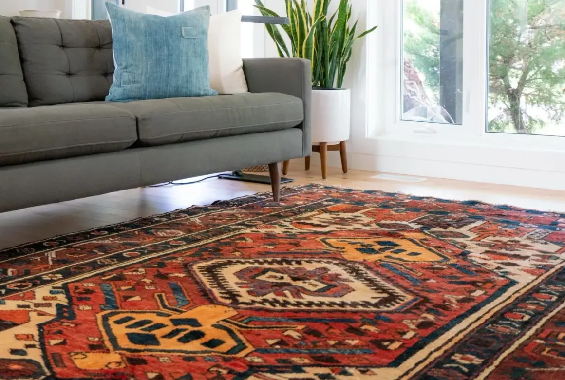 Ein detailreicher perserteppich mit roten und blauen Mustern liegt vor einem grauen Sofa. Tipps zum perserteppich reinigen.