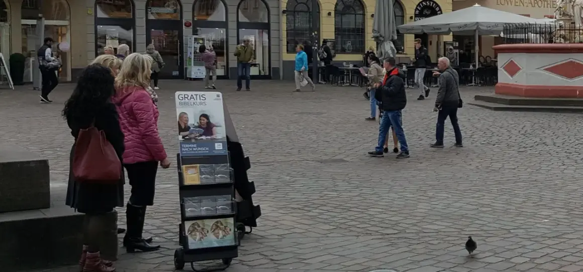 Zeugen Jehovas bieten einen kostenlosen Bibelkurs an. Menschen gehen vorbei, einige halten Broschüren in der Hand.