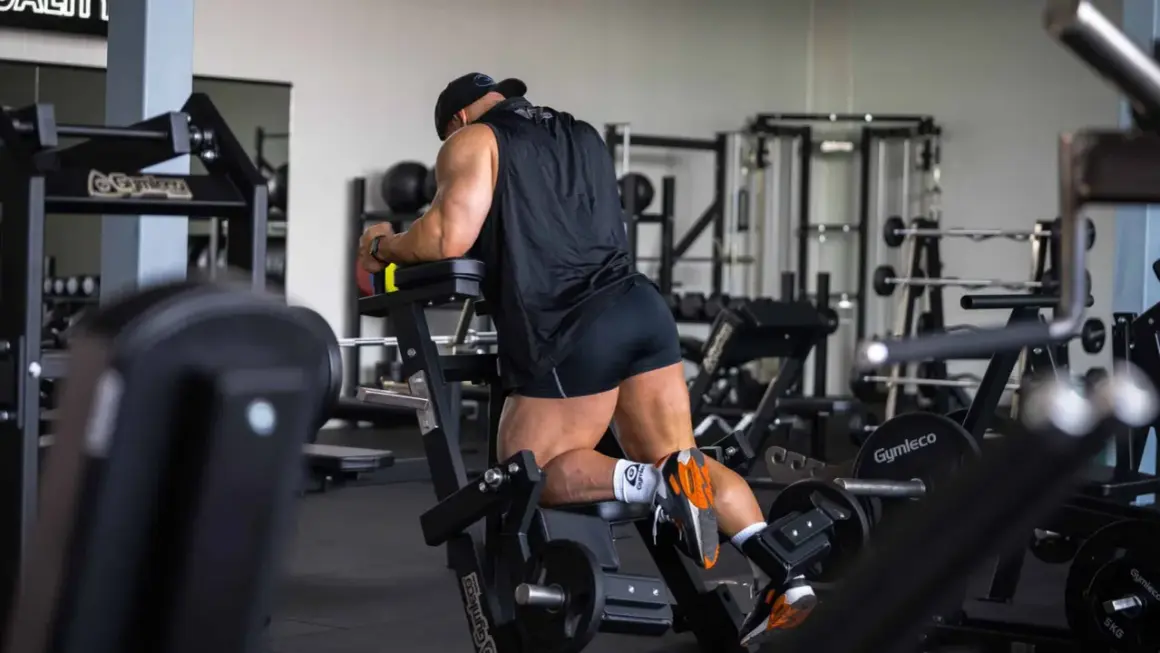 Man utför stående lårcurl på ett gym. Han har en svart tröja och shorts, och tränar sina baksida lår.